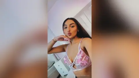 Young woman in a pink floral lace bralette posing confidently in a bright, modern bedroom setting.