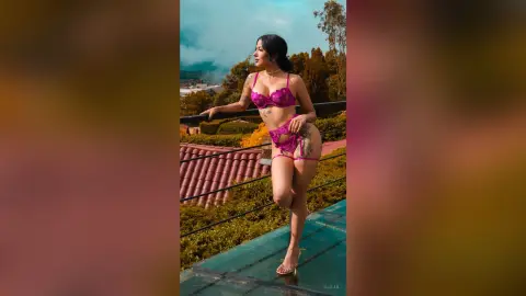 Aleja poses outdoors in pink lace lingerie, standing on a balcony overlooking a scenic landscape.