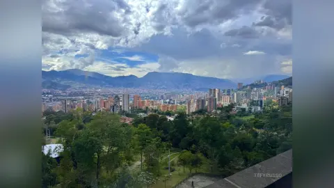 TefyRusso overlooking a wide view of a dense city valley surrounded by green trees and large mountains under a cloudy sky.