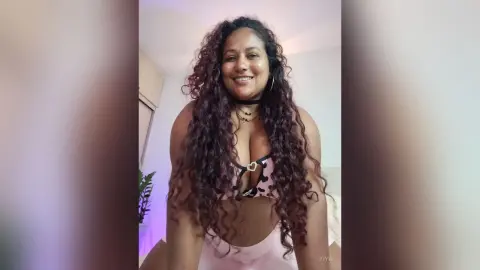 Smiling woman with long curly hair wearing a black and pink patterned bikini in a bright indoor setting.