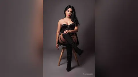 Glamorous woman in a black corset and thigh-high boots sitting on a chair against a grey studio background.