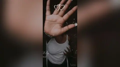 Young woman in a white tank top making a heart shape with her hands against a dark background.