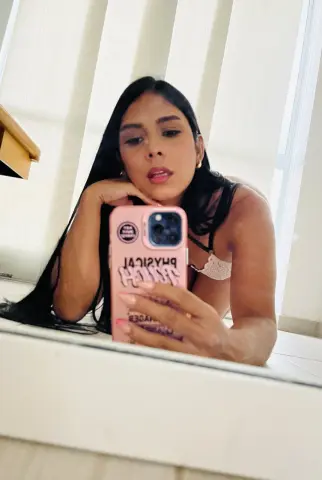 Young woman with long dark hair taking a relaxed selfie in white lace lingerie by a bright window.