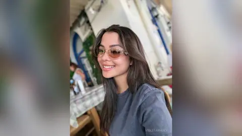 Smiling young woman with long brown hair wearing sunglasses and a grey t-shirt in a bright cafe setting.