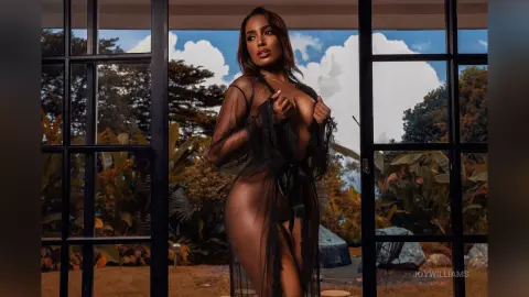 Confident woman in a black sheer lace robe standing by a window overlooking a lush tropical garden.