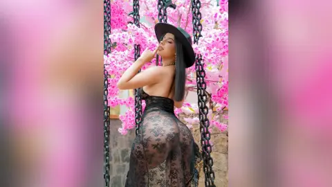 Young woman in a black lace outfit and wide-brimmed hat posing playfully amidst blooming pink cherry blossoms.