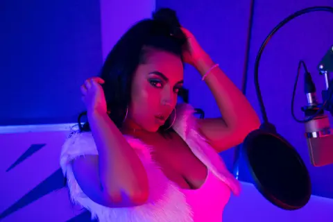 Glamorous woman in a white faux fur vest posing in a moody, neon blue and purple lit recording studio.