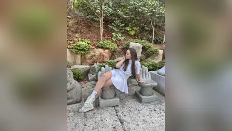 Young woman in a white summer dress sitting on stone sculptures in a lush green garden setting.