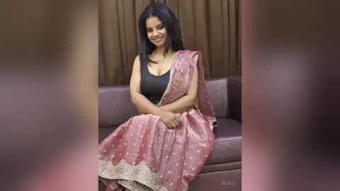 Smiling woman in a pink silk saree sitting on a sofa in a cozy indoor setting.