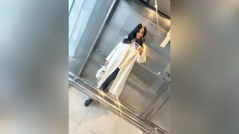 Stylish woman in a long white coat taking a mirror selfie in a modern elevator.