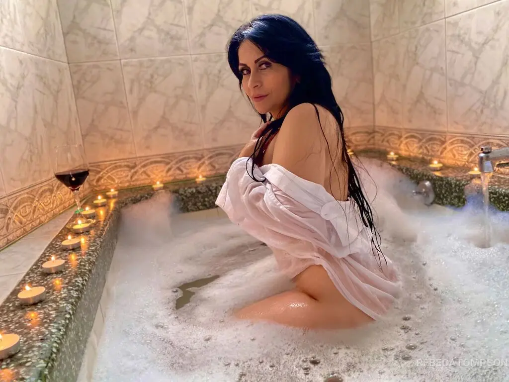 Relaxed woman in a white robe relaxing in a bubble bath surrounded by glowing candles in a marble bathroom.