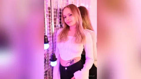 LucyLoy poses in a white long-sleeve crop top and black pants, showing off her stomach tattoo against a sparkly pink background.