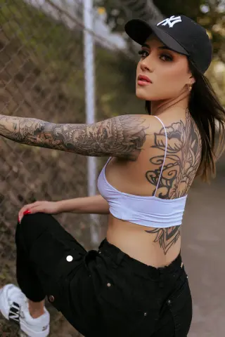 Tattooed woman in a white crop top and black pants posing by a chain-link fence in a casual urban setting.
