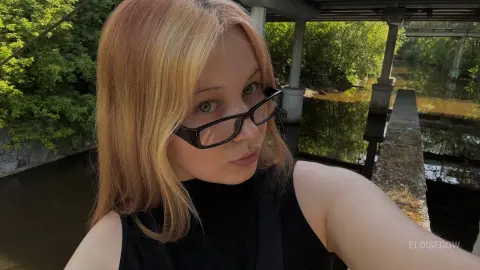EloiseGow wearing black glasses and a black top, posing outdoors near a river and bridge.