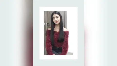 Young woman with long dark hair wearing a burgundy off-the-shoulder fuzzy sweater in a soft indoor setting.