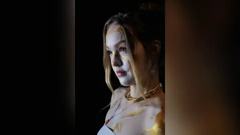 AliceTucci poses with gold and white paint or makeup covering her face, neck, and chest, wearing a gold choker against a black background.