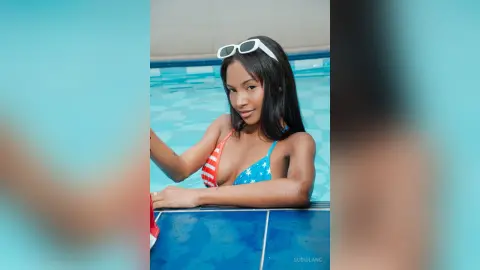 Smiling young woman in a red, white, and blue bikini relaxing by a bright blue swimming pool.