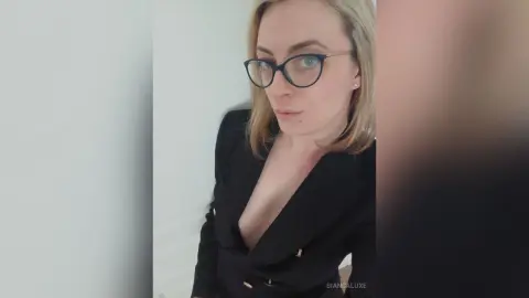 Confident blonde woman in a black blazer and glasses posing against a white wall.