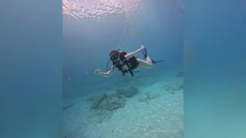 Female scuba diver in black gear swimming through clear blue ocean water over a sandy seabed.