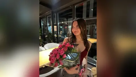 Smiling young woman in a red top holding a large bouquet of pink roses in a modern restaurant setting.