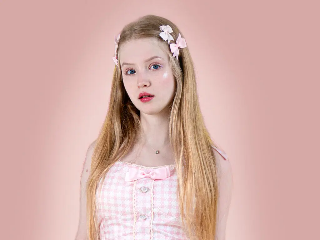 Blonde girl in pink gingham dress with hair bows posing against a soft pink background in a dreamy aesthetic.