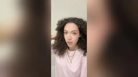 Young woman with curly dark hair wearing a white t-shirt and gold necklaces in a bright minimalist setting.
