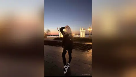 Blonde woman in black casual outfit standing on a stone walkway during a golden sunset by the water.