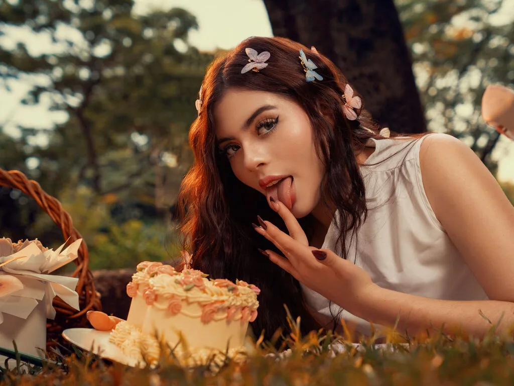 RachelRouge licks her lips while lying in the grass next to a cake, wearing a white sleeveless top with her hair decorated with small flowers.