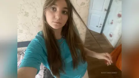 MartineKibbey with long brown hair wearing a bright blue t-shirt, looking at the camera in a room with floral wallpaper.