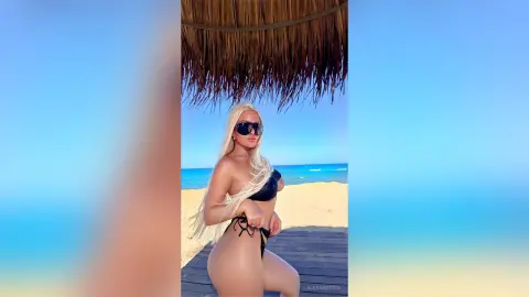 Blonde woman in a black bikini relaxing under a straw umbrella at a sunny tropical beach.