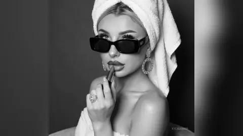 Glamorous woman in a white towel and sunglasses applying lipstick in a moody black and white portrait.