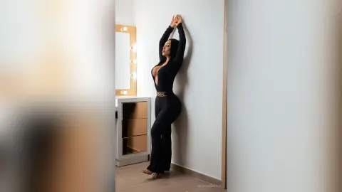Elegant woman in a black long-sleeve jumpsuit posing against a white wall in a modern dressing room.