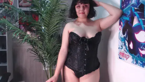 Smiling woman in a black lace corset standing near a green houseplant in a bright room.
