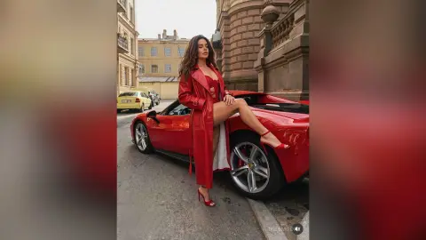 InesMoore poses outside a red sports car wearing a long red coat and red high heels, showing off her long brown hair and legs.