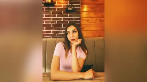 Thoughtful woman in a pink t-shirt sitting at a cafe table against a rustic brick wall background.