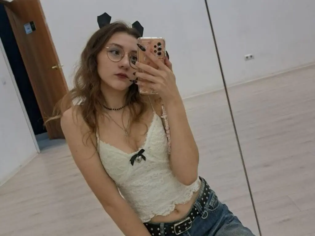 Young woman with glasses in a white lace camisole and jeans taking a mirror selfie in a bright room.