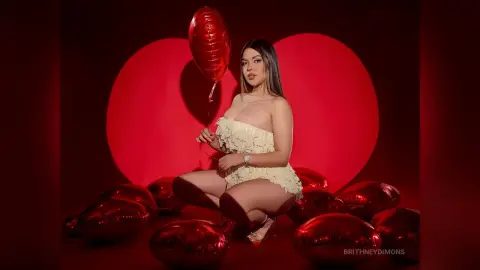 BrithneyDimons poses in a white lace strapless outfit, surrounded by red heart-shaped balloons against a red background.