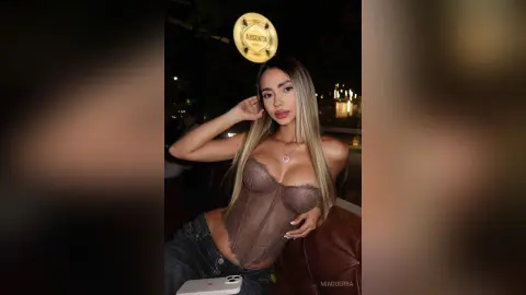 MiaGuerra poses in a brown lace corset and denim jeans, showing cleavage with her long blonde hair down.