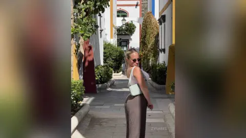 Stylish woman in a white tank top and brown trousers walking through a sunny Mediterranean village alleyway.