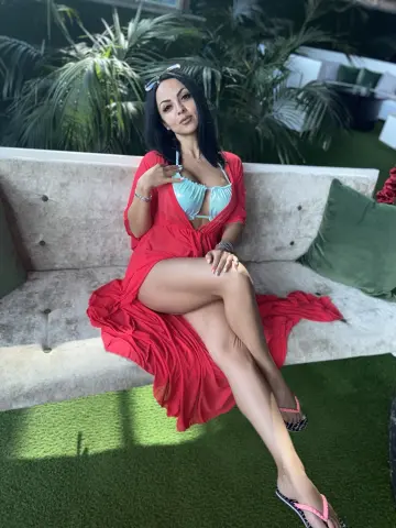 Issadorra sits on a white sofa wearing a light blue bikini top and a long red dress that is open to show her legs, surrounded by green palm leaves.