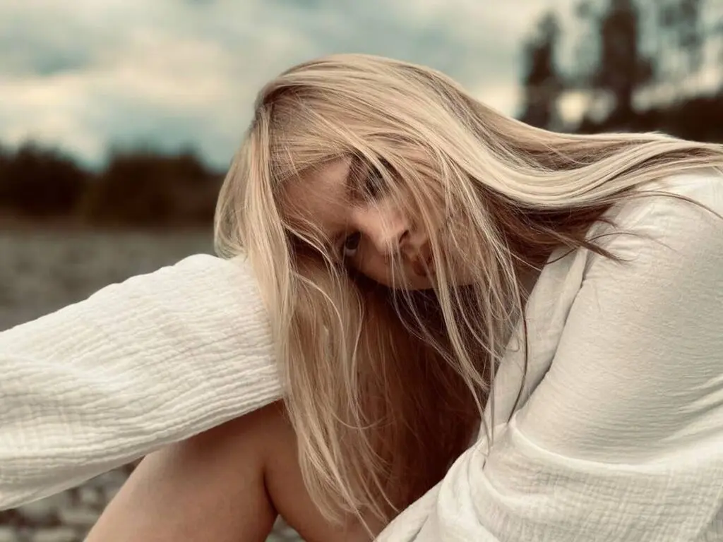 Blonde woman in a white textured shirt relaxing outdoors in a moody, natural setting.