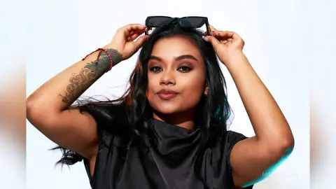 Smiling woman with long dark hair and tattoos wearing a black sleeveless top against a white background.