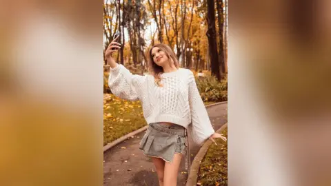 Smiling woman in a white cable knit sweater and denim skirt taking a selfie in an autumn forest setting.