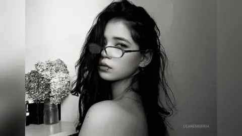 DeaneMerrih poses with long, dark wavy hair and glasses, looking over her shoulder with bare shoulders against a plain wall.