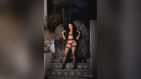 Mystical woman in black lace lingerie and large feathered wings standing on a patterned stone staircase at night.