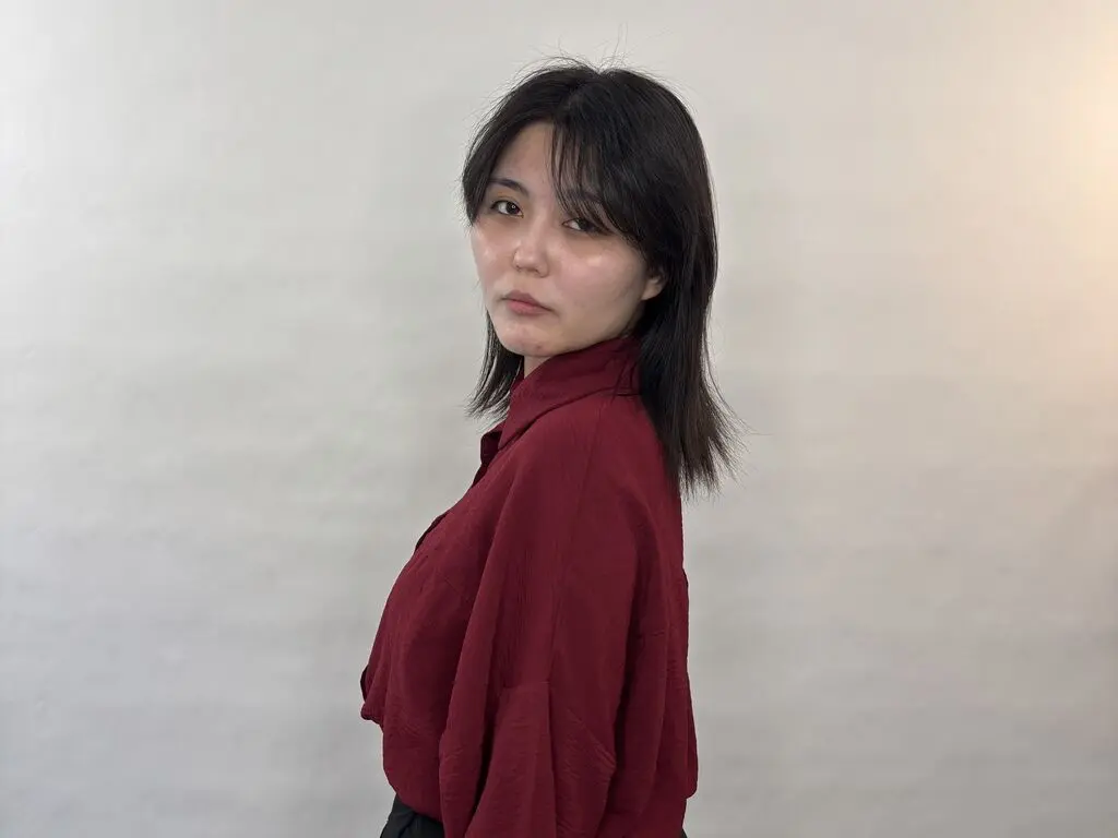 Young woman with dark hair wearing a burgundy button-down shirt against a minimalist white background.