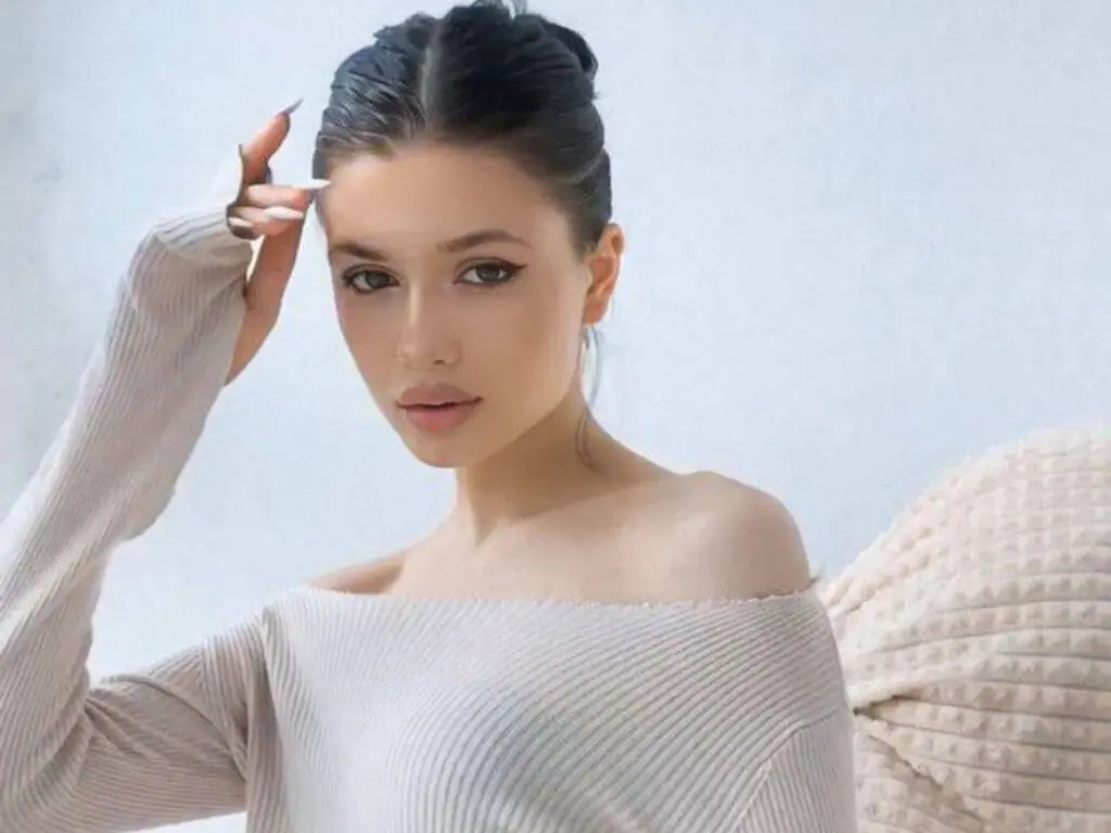 Young woman in a beige ribbed off-the-shoulder sweater posing with a serene expression against a light background.