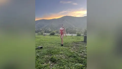 Woman in a red one-piece swimsuit standing in a grassy field against a scenic mountain sunset backdrop.