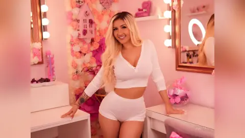 Anita smiles while wearing a white long-sleeve crop top and matching white shorts in a pink-themed room.