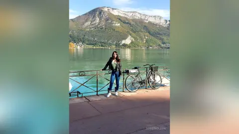RossiNoleen stands by a metal railing overlooking a calm lake and mountains, wearing a black top and blue jeans.